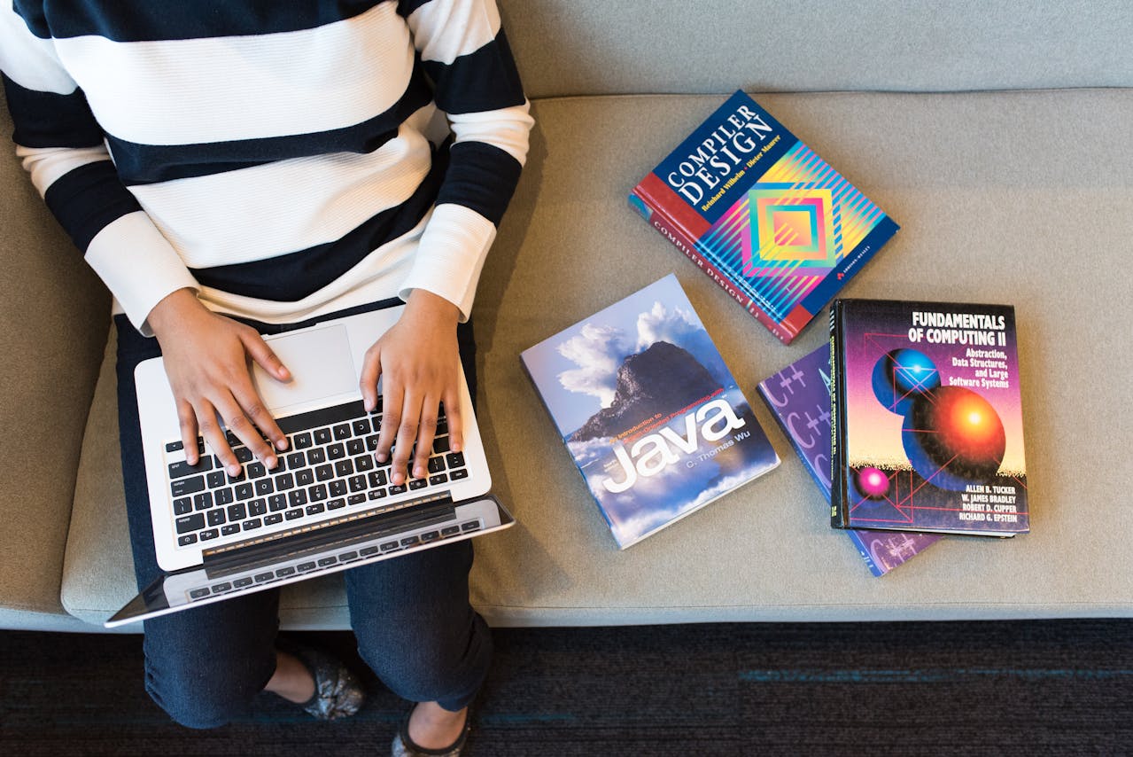 A woman sitting on a grey couch with four coding books next to her with one of them being for Java to depict the use of Java in 2025 Image source: https://www.pexels.com/photo/person-using-macbook-pro-on-person-s-lap-1181298/