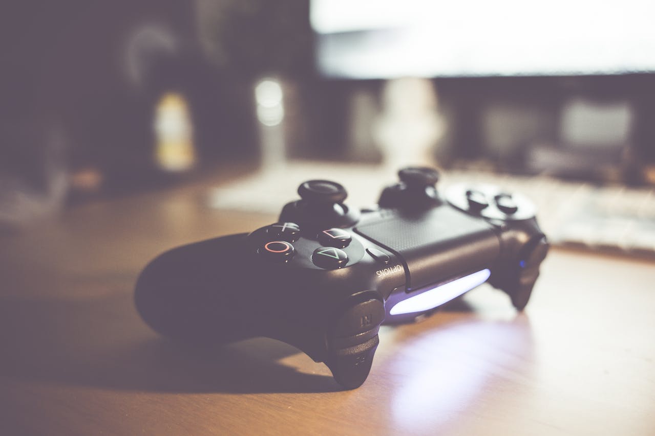 a picture of a black PS5 controller sitting on a desk with a monitor behind it to represent one of the consoles on which you can play Assassin's Creed Shadows once it comes out image source: https://www.pexels.com/photo/black-sony-ps4-controller-539986/